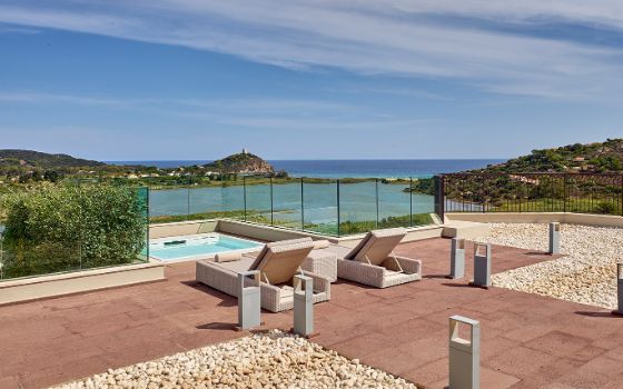 Zwei Sonnenliegen an einem Jacuzzi auf einer Hotelterrasse an einer mediterranen Küstenlandschaft unter blauem Himmel.