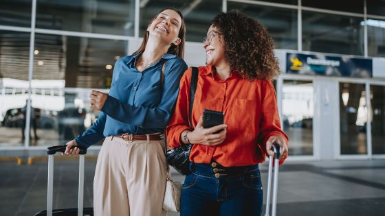 Zwei elegant gekleidete Frauen mit Trolleys stehen lachend vor dem Eingang eines Flughafenterminals.