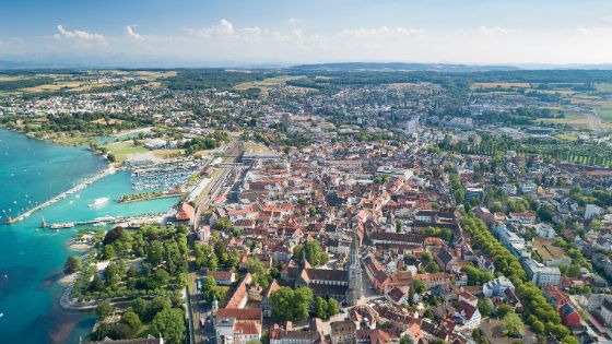 Luftaufnahme von Konstanz am Bodensee mit Altstadt, Hafen, Bahnhofsbereich und umgebender Landschaft an einem sonnigen Tag.