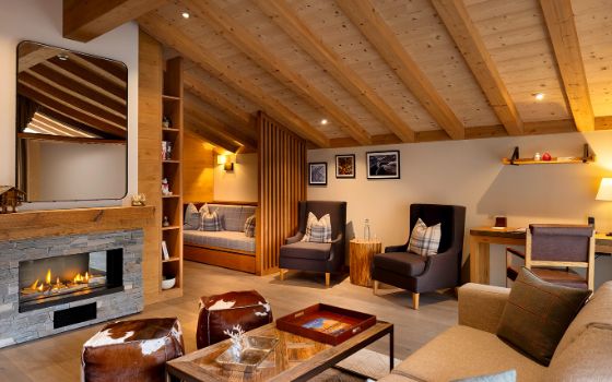 In einem hellen, grossen Raum mit Holzdecke befindet sich ein kleiner Kamin mit Tisch, Hockern und Sofa. Im hinteren Bereich des Raumes stehen zwei Sessel und ein Schreibtisch mit Stuhl an der Wand. 