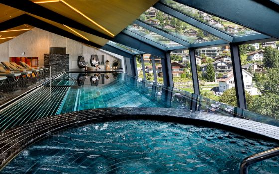 Zwei Personen in Hängesesseln an einem modernen Indoor-Pool mit Jacuzzi unter einem Panoramadach mit Blick auf eine Ortschaft an einem Berghang.