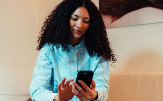 Eine sitzende Frau bedient ein Smartphone in ihrer Hand vor einer Wand mit Gemälde.