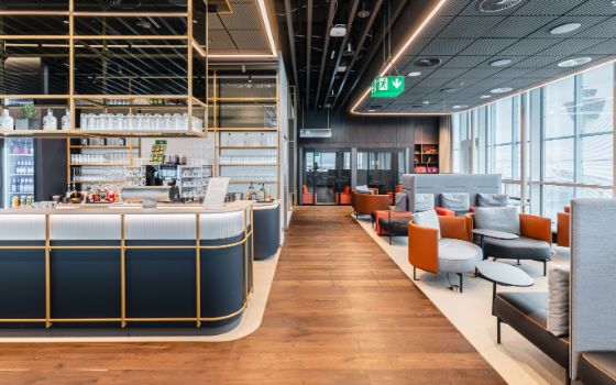 Raum einer Lounge am Flughafen München mit Holzboden, Sofas, Sesseln, Fensterfront zum Rollfeld und einer Bar.
