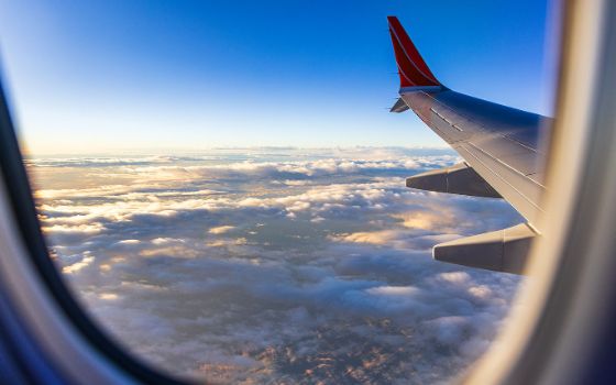 Blick aus einem Flugzeugfenster auf eine Tragfläche vor blauem Himmel über einer wolkenverhangenen Landschaft