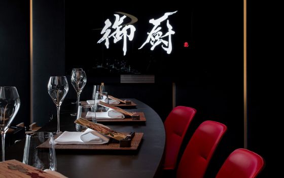 Rote Stühle an einem edel eingedeckten Tresen in einem schwarzen Raum mit weissen japanischen Schriftzeichen an der Wand. 