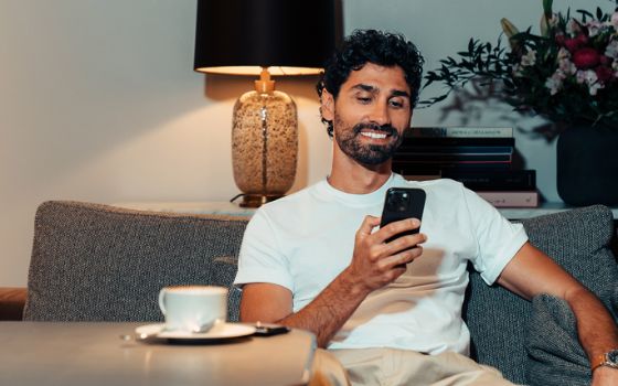 Ein Mann im weissen T-Shirt sitzt entspannt auf einem Sofa an einem Tisch mit Kaffeetasse und schaut lächelnd auf ein Smartphone in seiner Hand.