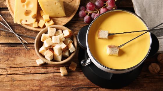 Ein Topf mit Käsefondue steht auf einem Holztisch, darum sind Brotstücke, Weintrauben und Käse drapiert.