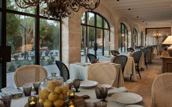 In einem Restaurant mit hohen Fenstern und hellen Fliesen stehen gedeckte Tische mit Leinendecken und Korbstühlen. 