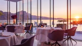 In einem Restaurant stehen gedeckte Tische und Stühle. Durch die bodentiefen Fenster blickt man auf den Vierwaldstättersee und Berge. 