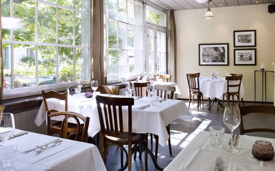 Eleganter, heller Speisesaal im Brasserie-Stil mit weiss eingedeckten Tischen und Holzstühlen.