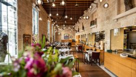 Elegant eingedeckte Tische in einem modernen Restaurant Restaurant mit Holzboden und Steinmauern mit Panoramafenstern, von der Decke hängen zahlreiche runde Glaslampen.
