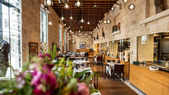 Elegant eingedeckte Tische in einem modernen Restaurant Restaurant mit Holzboden und Steinmauern mit Panoramafenstern, von der Decke hängen zahlreiche runde Glaslampen.