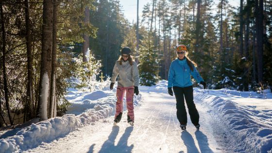 Zwei lachende Frauen auf Schlittschuhen in einem verschneiten Wald. 
