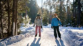Zwei lachende Frauen auf Schlittschuhen in einem verschneiten Wald. 