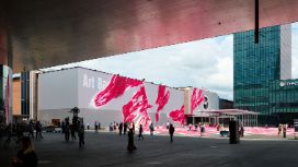 Personen auf einem Platz mit Gebäuden der Art Basel mit von moderner Kunst verzierten Fassaden. 