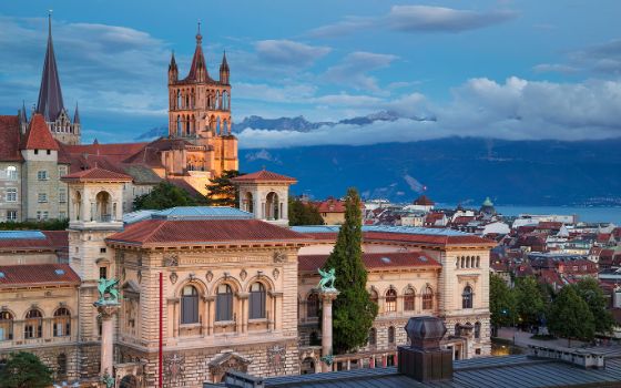 Blick auf die Kathedrale von Lausanne mit ihrem markanten Turm, dahinter die Stadt und der Genfersee mit Alpenpanorama.