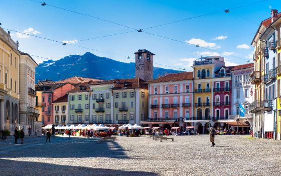 Marktplatz mit Verkaufsständen, umgeben von bunten Häusern vor Bergpanorama bei Sonnenschein. 