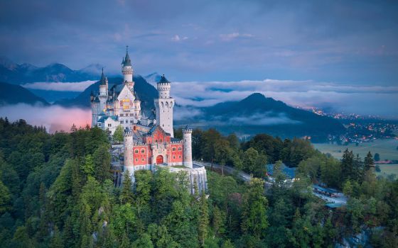 Märchenhaftes Schloss Neuschwanstein im Stil der Neuromantik auf einem bewaldeten Hügel vor mit Nebelschwaden verhangener Berglandschaft im Abendlicht. 