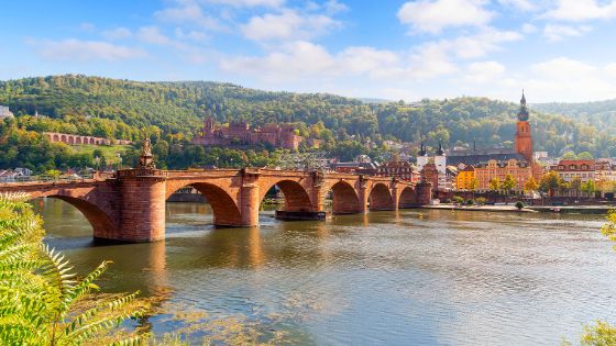 Stadtpanorama von Heidelberg am Fluss hinter einer Bogenbrücke aus rotem Sandstein.
