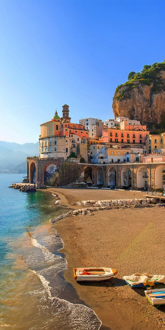 Mediterraner Fischerort an einer Felsküste mit bunten Häusern und Bogenbrücke entlang einer Bucht mit Sandstrand.
