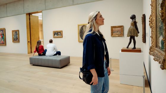 Eine Frau betrachtet ein Gemälde in einem verzierten, goldfarbenen Rahmen in einem Ausstellungsraum mit weiteren Personen und Kunstwerken.