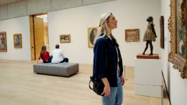 Eine Frau betrachtet ein Gemälde in einem verzierten, goldfarbenen Rahmen in einem Ausstellungsraum mit weiteren Personen und Kunstwerken.