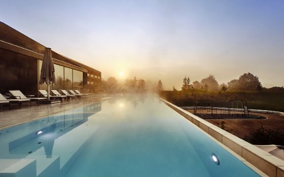 Ein Hotel Aussenpool mit Liegen ist umgeben von Natur, im Hintergrund geht die Sonne auf.