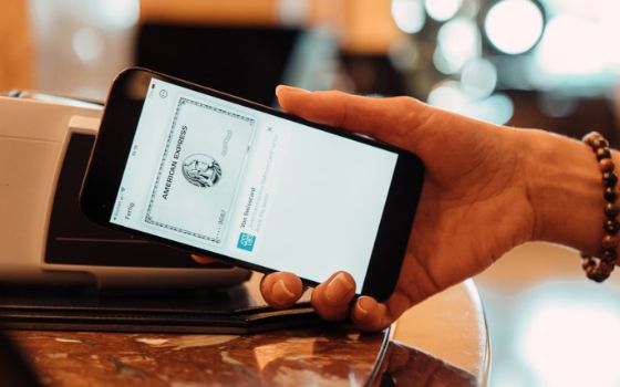 Nahaufnahme einer Hand, die ein Handy an die Seite eines Kartenlesegerätes hält, auf dem Display eine American Express Platinum Card