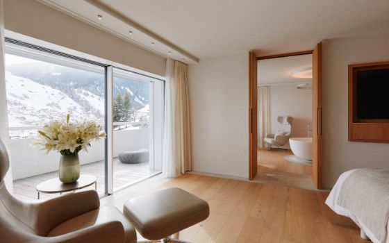 Eine Suite in einem Hotel mit einem Ledersessel und Bett in einem Raum, einer offenen Tür zu einem Badezimmer mit Badewanne und einem grossen Balkon, im Hintergrund verschneite Berge.