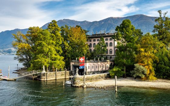 Luxuriöse Villa im neoklassizistischen Stil mit Bootsanleger und Schweizer Flagge auf einer Insel in einem See vor Bergpanorama. 