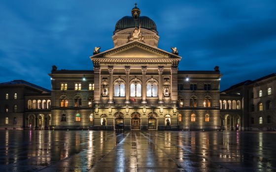 Das beleuchtete Bundeshaus in Bern bei Abenddämmerung mit nasser Spiegelung des Bundesplatzes im Vordergrund.