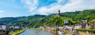 Mittelalterliche Burg auf einem grünen Hügel oberhalb einer Kleinstadt mit Fachwerkhäusern an einem Fluss mit Ausflugsbooten.