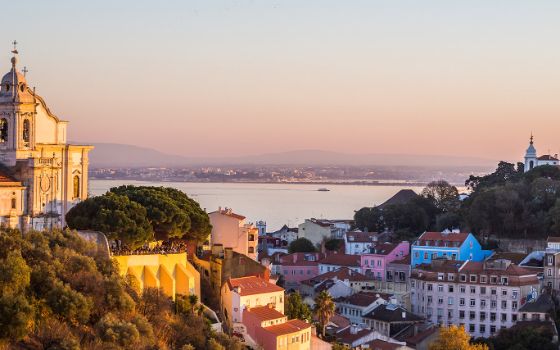 Panoramablick auf Lissabon bei Sonnenuntergang mit pastellfarbenen Häusern, Tejo und Kirchenfassade im Vordergrund