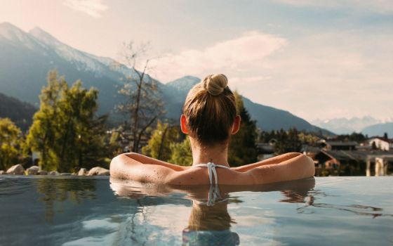 Rückansicht einer Frau in Badekleidung in einem Outdoor-Pool; sie steht mit aufgestützten Armen am Beckenrand vor einer Berglandschaft. 