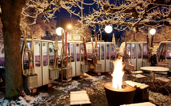 Weisse Skigondeln stehen Reihe an Reihe auf einem Platz, vor dem ein Lagerfeuer brennt. 