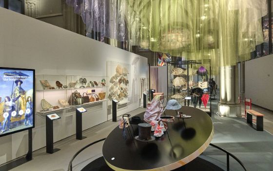 In einem hellen Raum sind verschiedene Accessoires ausgestellt wie Taschen, Hüte, Schuhe und Fächer, von der Decke hängen glitzernde Stoffe. 