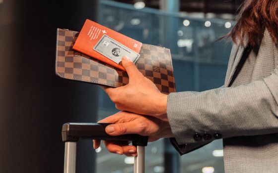 Eine silberne Kreditkarte von American Express auf rotem Reisepass und Brieftasche in der Hand einer Frau über dem Griff eines Koffers in der anderen Hand in einem Flughafenterminal 