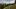 Eine Frau und zwei Kinder laufen auf einem Weg zwischen Wiesen sowie Bäumen und mit Bergpanorama. 