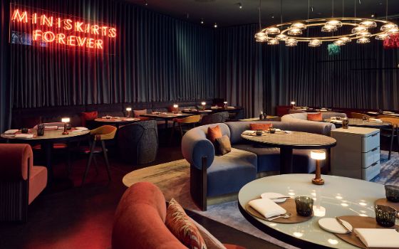 Moderner, stilvoller Speisesaal mit einer Neonschrift vor blauen Vorhängen und geschwungenen Designsesseln mit Samtbezug an runden, eingedeckten Tischen.
