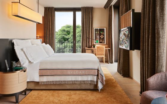 Elegantes Hotelzimmer mit Textilien in warmen Erdtönen und Doppelbett mit einer Wolldecke mit dem Logo von Bulgari, dahinter ein Fenster mit Blick ins Grüne. 