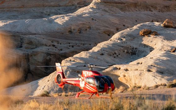 Ein roter Hubschrauber steht auf einem sandigen Boden vor einer roten Felswand.