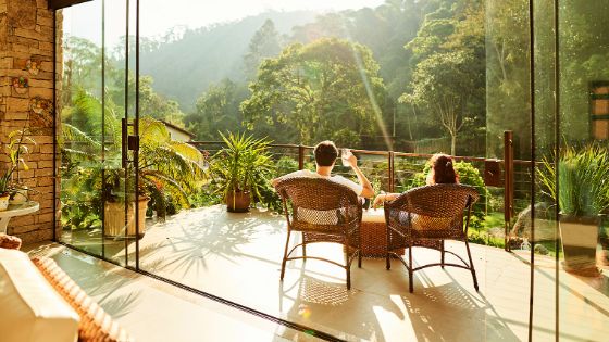 Rückansicht eines Paares mit Getränken in Rattansesseln auf einem grossen Balkon vor grüner Dschungellandschaft bei Sonnenschein