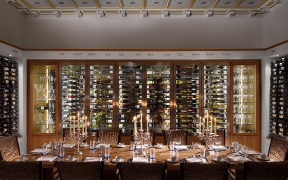 Vor einem grossen gläsernen Wandschrank mit etlichen Weinflaschen steht ein langer gedeckter Tisch, an der Wand sind weitere Weinregale.