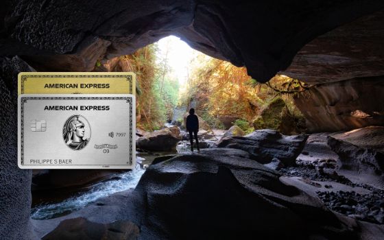 Eine Person steht in einer Höhle und schaut auf eine idyllische Waldlandschaft. Im Vordergrund sieht man eine platinfarbene und eine goldene Kreditkarte von American Express.