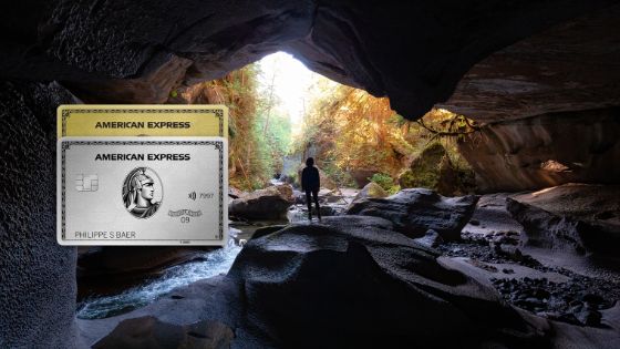 Eine Person steht in einer Höhle und schaut auf eine idyllische Waldlandschaft. Im Vordergrund sieht man eine platinfarbene und eine goldene Kreditkarte von American Express.