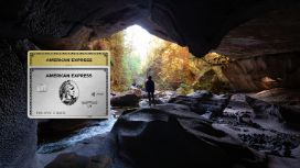 Eine Person steht in einer Höhle und schaut auf eine idyllische Waldlandschaft. Im Vordergrund sieht man eine platinfarbene und eine goldene Kreditkarte von American Express.