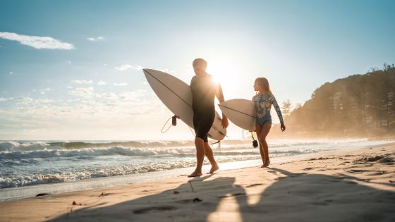 Zwei Personen in Surfkleidung mit Surfbrettern unter dem Arm laufen in der Abendsonne an einem Strand am Meer entlang.