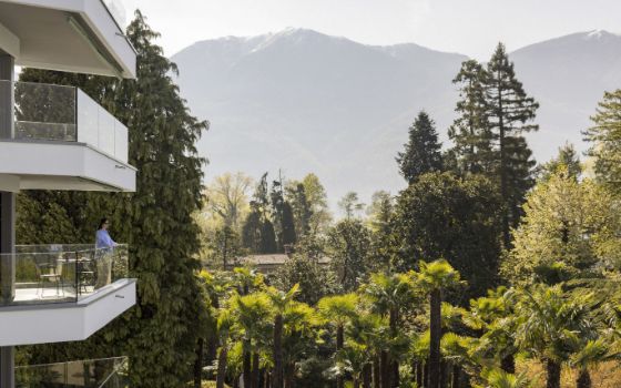Eine Person steht auf einem Balkon und blickt in die Ferne, im Hintergrund ein Bergpanorama und Bäume.