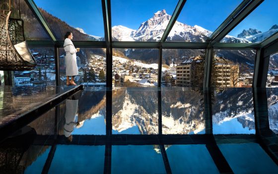 Eine Frau im weissen Bademantel steht an einem Panoramafenster an einem Innenpool, in dem sich die verschneite Berglandschaft des Hintergrundes spiegelt.