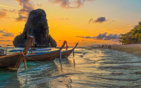 Mehrere Langheckboote vor einer Felsformation im flachen Wasser an einem Sandstrand mit Personen bei Sonnenuntergang. 